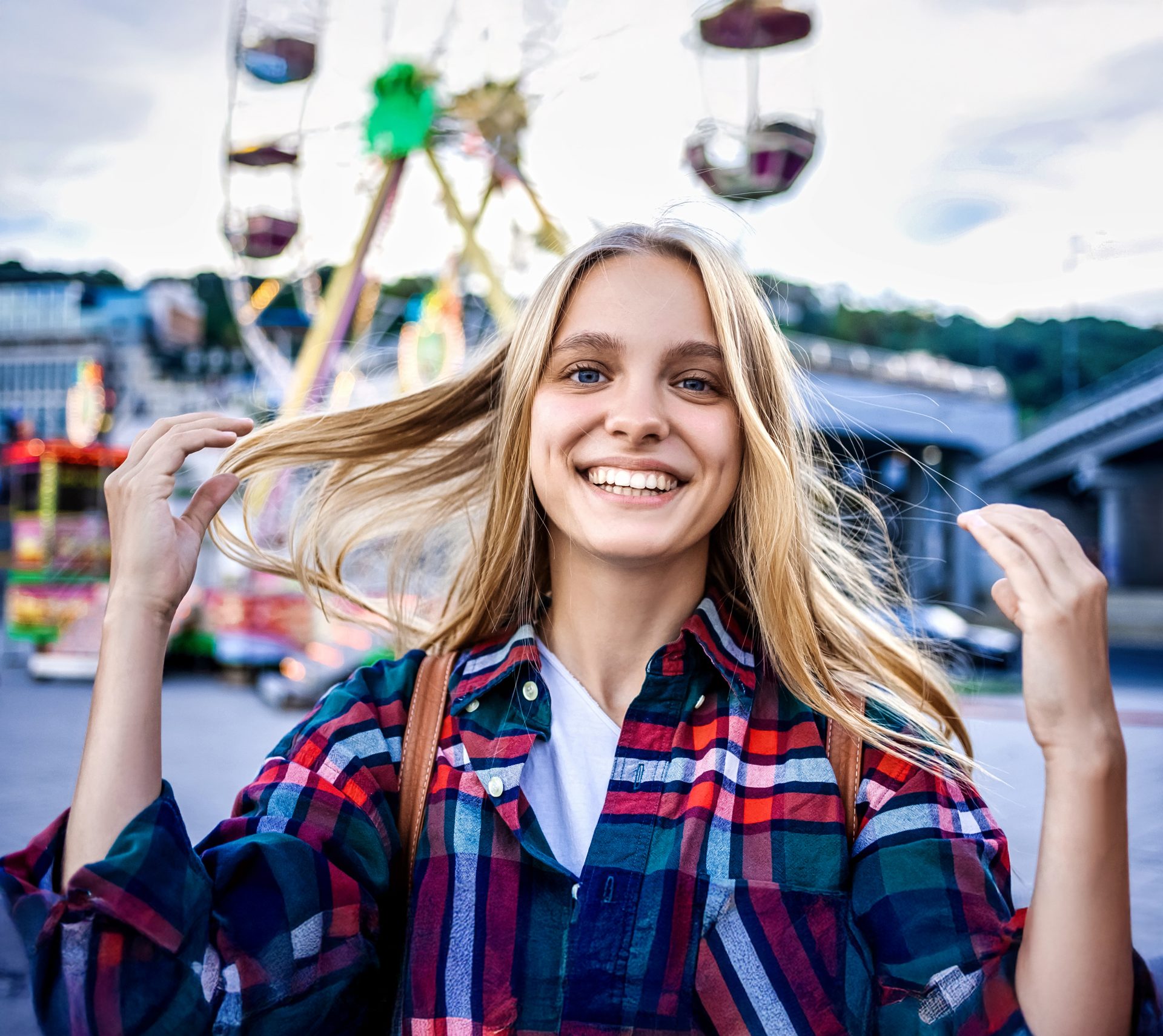 Uśmiechnięta młoda kobieta z blond włosami na tle wesołego miasteczka, prezentująca zdrowe zęby – przykład efektu leczenia ortodontycznego z użyciem nakładek Invisalign®.