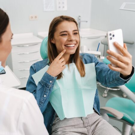 Młoda kobieta w gabinecie stomatologicznym uśmiecha się i robi selfie, wskazując na swoje zęby. Obok siedzi dentystka w maseczce. Scena związana z konsultacją ortodontyczną i leczeniem nakładkami Invisalign®.
