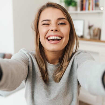 Młoda kobieta w szarej bluzie uśmiecha się szeroko i mruga, robiąc selfie w jasnym wnętrzu. Zdjęcie ilustruje naturalny uśmiech możliwy do osiągnięcia dzięki leczeniu nakładkami Invisalign® w CHEESE.
