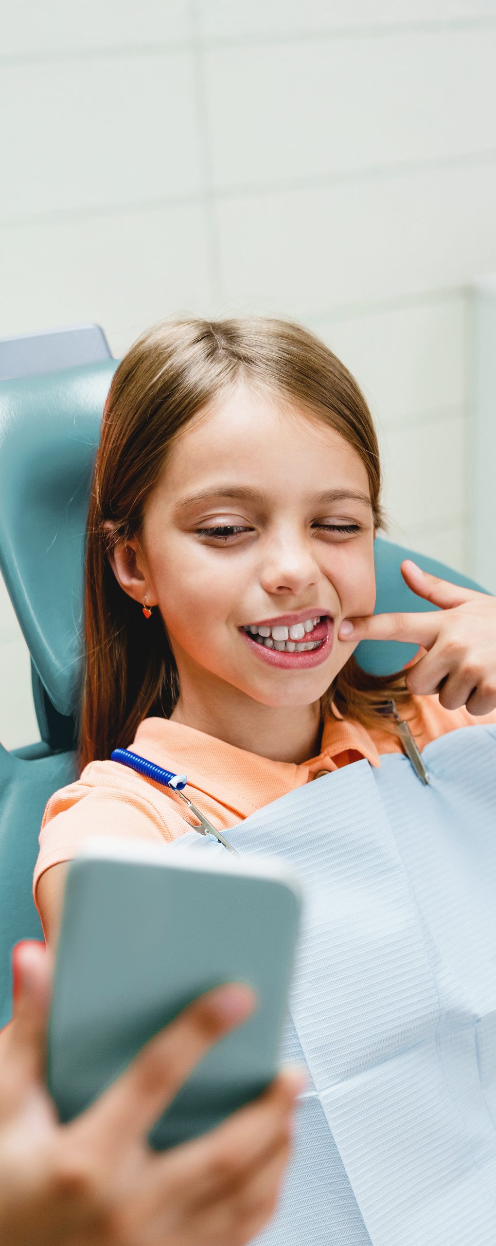 Dziewczynka na fotelu dentystycznym, uśmiecha się i wskazuje na swoje zęby podczas robienia zdjęcia smartfonem – przykład wizyty ortodontycznej i diagnostyki przed leczeniem nakładkami Invisalign®.
