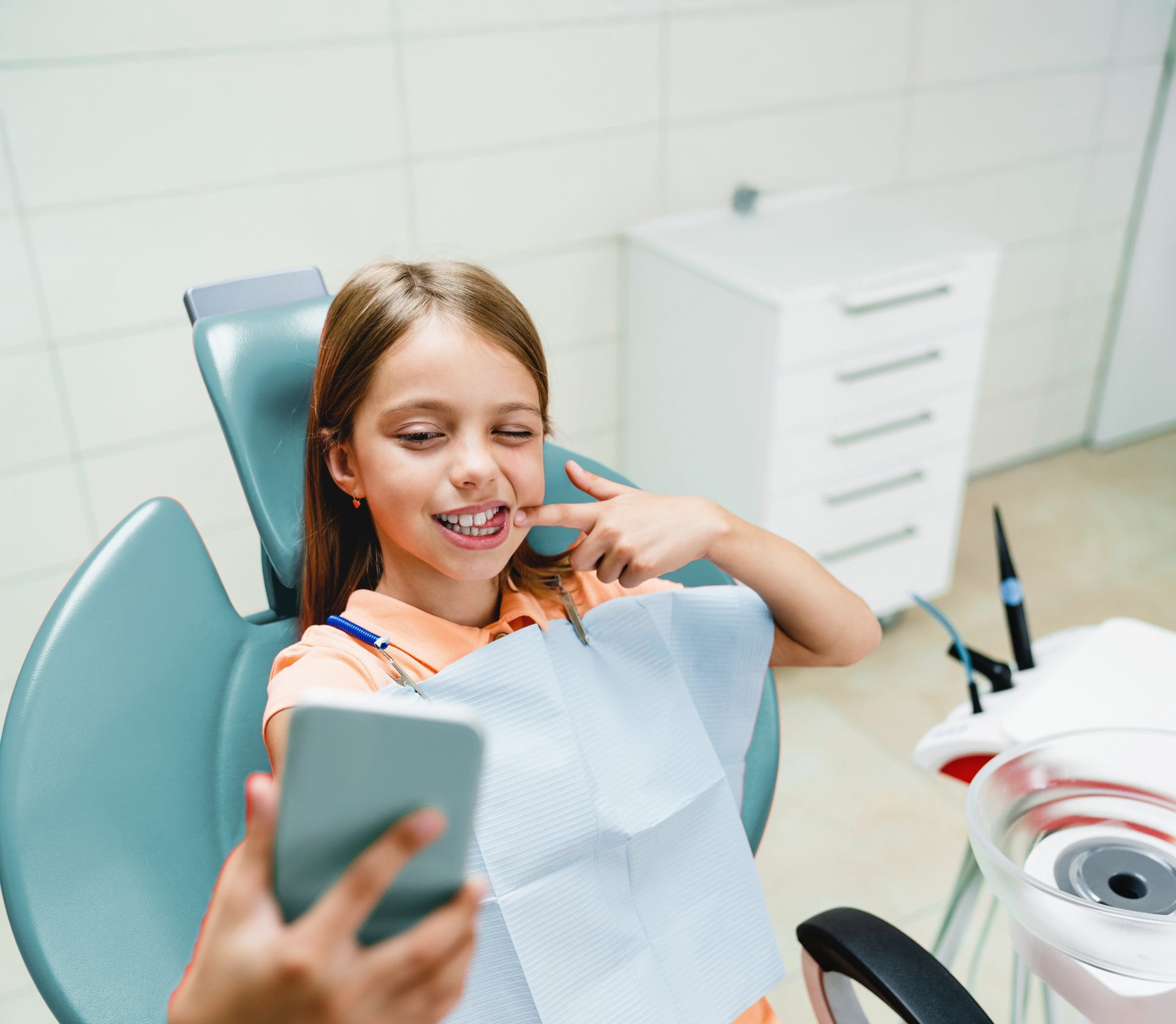 Dziewczynka na fotelu dentystycznym, uśmiecha się i wskazuje na swoje zęby podczas robienia zdjęcia smartfonem – przykład wizyty ortodontycznej i diagnostyki przed leczeniem nakładkami Invisalign®.