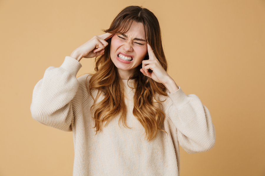 Młoda kobieta z wyrazem silnego bólu na twarzy, trzymająca się za skronie na jasnobrązowym tle.